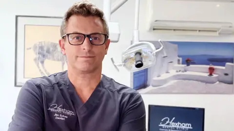 Photo of dentist Jim, a middle-aged man with fair hair and black-rimmed glasses. He's wearing scrubs and standing in a dental treatment room.