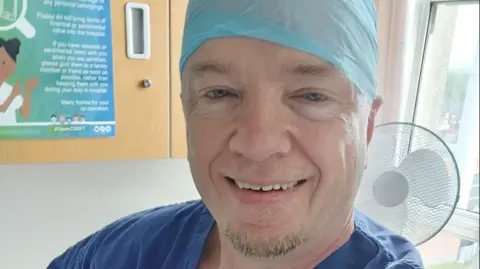 Instagram | Robert Charles Albon Joe Albon with blue eyes smiling at the camera. He is wearing blue hospital scrubs and a light blue protective covering over his head. 