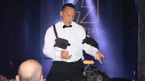 Piers Burnell is wearing evening dress, minus the blazer, whilst air guitaring in front of a crowd.