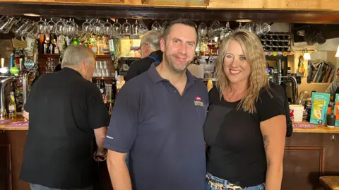 Pub owners Steve, wearing blue T-shirt, and Abi Dean in black top. Both are stood in front of a wooden bar with glasses hanging above.