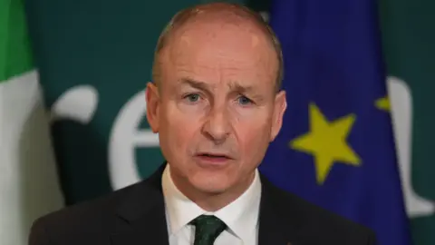 PA Media A man with light hair, he is wearing a navy blazer, white shirt and green tie with shamrocks