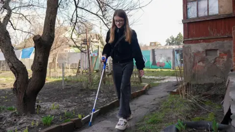 Lera walks by her house using one crutch