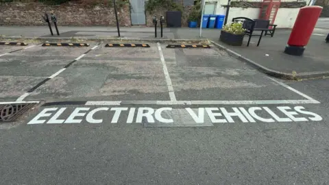 Two electric vehicle bays with misspelt road markings reading "electirc vehicles" at the side of a residential street. A traditional British phonebox and letterbox can be seen on the pavement.