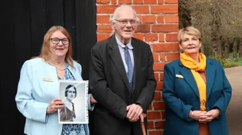 Jane Miles Sue MacEwen in a sky-blue blazer holds a photo of Lady Petre. She has ginger hair and glasses. Centre is Lord Petre in a pinstripe black suit. To the right is Moyra Jackson in an orange scarf and blue blazer. Behind them is a bricked archway.