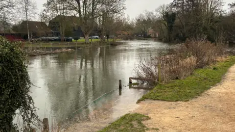 Buildings including a barn stand behind a river which has burst its banks. A sandy path is in the corner of the photo.