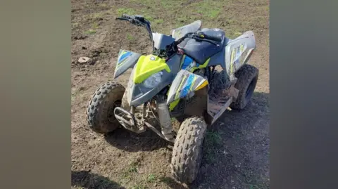 A quad bike parked on uneven, muddy ground. Dirt and dried mud are visible on the bike and tyres.