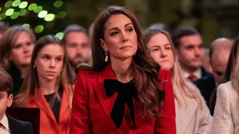 PA Media Catherine is pictured at a previous carol event. She is wearing a red jacket and has brown hair. She is sat in front of a number of people.
