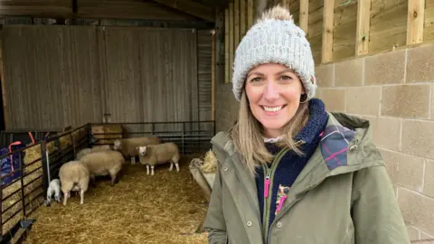 Hannah Tuckwell is standing in front of a barn. She is wearing a green coat and a light blue bobble hat. Behind her, on a bed of straw, a number of sheep and lambs are standing.