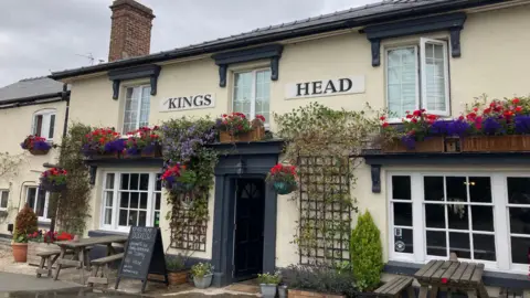 The Kings Head is painted cream with dark navy blue doorways and window lintels. The windows are white and there are plants and vines growing up the side of the building. 