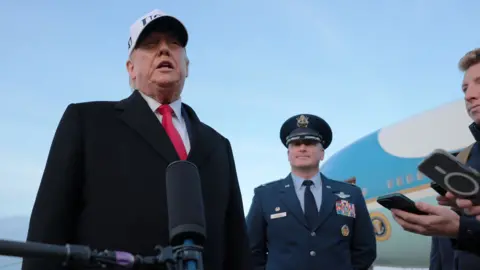 Donald Trump wears a white cap saying 'USA' and a suit and red tie as he speaks into a microphone next to a military officer and a reporter recording him with their phone, on the tarmac next to Air Force One against a blue sky in Maryland on 13 January.