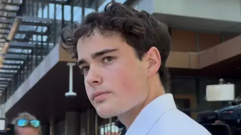 A young man with brown hair and a white shirt looks at the camera