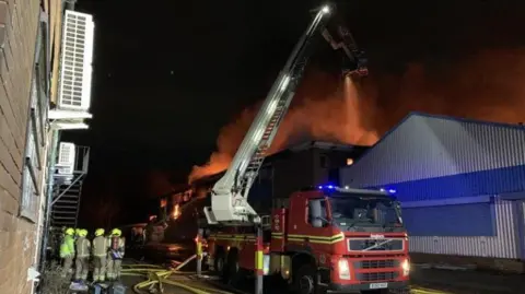 West Midlands Fire Service Smoke and flames rise up from a large building, with a fire engine and firefighters in the foreground