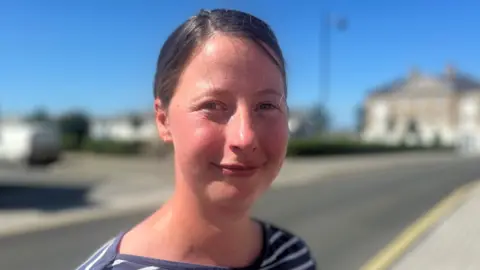 A young woman with brown hair tied back in a ponytail looking straight at the camera. She is wearing a blue and white striped top. The sky is blue and she is standing by a road.