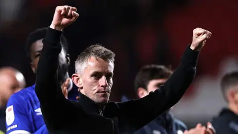 Getty Images A man raises both his arms in the air in celebration. He wears a black jumper. Football players wearing blue shirts can be seen clapping behind him. 