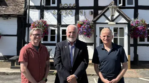 Three men are standing outside a pub called the Rose & Crown - one man is wearing a red shirt, the man in the centre is wearing a black suit jacket and a black and white striped shirt, and the third man is wearing a black top. 