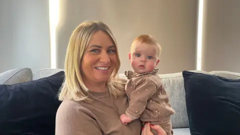 Holly Nichols/BBC The image shows Mrs Madden-Giles sitting on a light grey sofa holding her four month old daughter, Tulley. Both are wearing brown clothing, and there are two large dark cushions on the sofa. The background features tall, light-coloured blinds with narrow gaps letting in light.