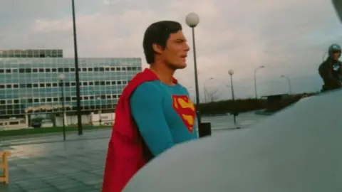 Stephanie English Christopher Reeve in a Superman costume stands outdoors in front of a modern building with many windows. The sky is cloudy and there are street lamps are in the background.