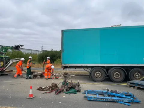 Workers in hi-vis clothing clearing the site. There are metal items laid out on the floor to the side of a blue trailer.