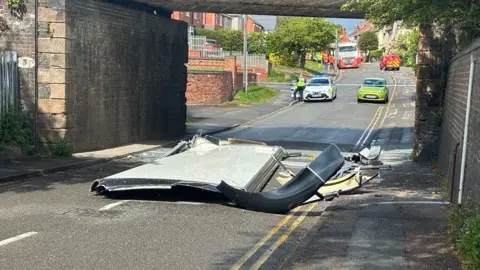 A bus roof under a bridge with debris strewn across the side of a road