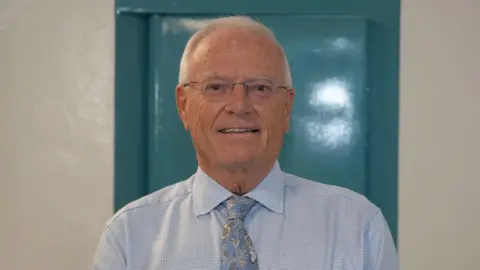 Declan Moore, the governor of HMP Norwich. He is standing in a white corridor and is looking directly at the camera and smiling. He is wearing a pale blue shirt with a blue patterned floral tie.