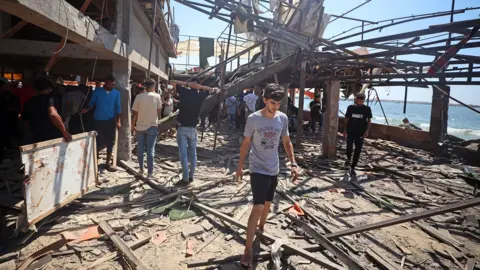 A view of the destruction after air strikes on a coffee shop in Gaza. People are pictured examining the debris with a young man walking out of the cafe in the foreground, and the sea and blue sky in the background, taken on 30 June.