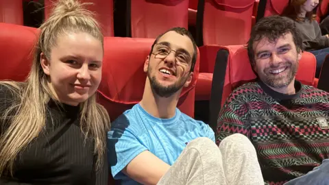 BBC Jimmy Simpson, who is wearing a blue shirt and light grey trousers and wearing thin-rimmed glasses, sits in between Lauryn Bell, who has blonde hair and is wearing a black shirt, and surf instructor Nick Jones who is wearing a striped-patterned jumper. They are all sitting on row of red cinema seats.