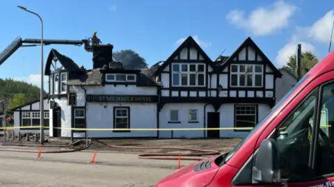 BBC Firefighters use an aerial platform to damp down a fire-damaged building labelled St Michaels Hotel, with part of the roof collapsed, hoses and a cordon in place, and a Scottish Fire and Rescue Service van in the foreground.