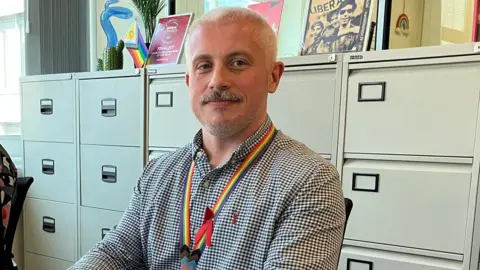 Ant Hopkinson has shirt hair and a moustache and beard. He is wearing a black and white checkered shirt and has a rainbow lanyard around his neck