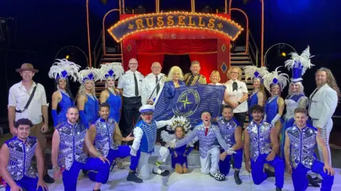 National Coastwatch Mablethorpe Circus performers dressed in blue and white outfits with ruffled white feather hats on the women holding the flag