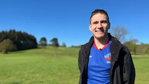 Kevin Oakes stands in Cofton Park, behind him is a vivid blue sky, trees and bright green grass. He has short hair and is wearing a blue running top and a black jacket.