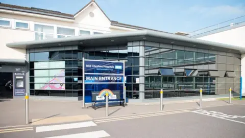 The main entrance to Leighton Hospital. A blue sign which says "main entrance" sits outside the front of a glass-built entrance building with the white hospital building visible behind it.