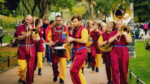 BCP Council A group of people dressed in bright red and yellow boiler suits while playing various brass, woodwind and percussion instruments and walking through Bournemouth