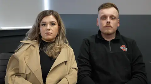 A man and woman sit together looking at the camera, the woman is wearing a camel coloured coat and her partner a black sweatshirt.