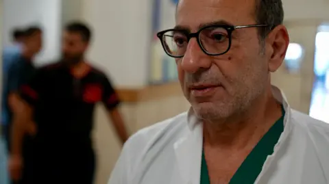 Dr. Goktay Koraltan Hassan Wazni poses for a photo in Nabih Berri government hospital. He is a middle-aged man wearing a white doctor's coat, green clothes, and black-rimmed glasses.