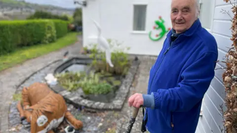 Malcolm Curley stands outside wearing a blue zip-up fleece jacket and holds a stick. Beside him is a large tiger sculpture lying down beside a smaller cat. In the background there's a bright green gekko on the wall and a pond with a bird in it. All are works of art.