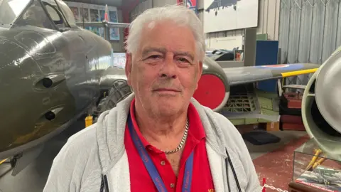 A man with white hair, wearing a red Midland Air Museum polo shirt, lanyard and grey zip-up jacket. He stands inside a hangar lined with historic military aircraft. The nose of a grey fighter jet sits to his left, with an RAF roundel on a wing behind him.