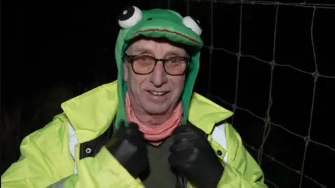 BBC A man wearing a felt hat in the shape of a frog and dark-rimmed glasses. He has a hi-vis jacket on and is wearing black leather gloves.
