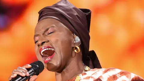 Angélique Kidjo sings into a microphone with her eyes closed. She wears gold hoop earrings, red lipstick and a brown headscarf.