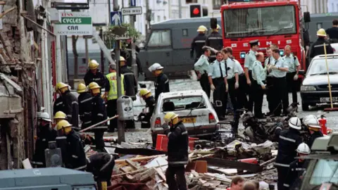 PA Firefighters and police officers attending to the aftermath of the Omagh Bomb. There is a white car amongst the wooden and metal rubble, with a fire engine, another black car and van in the background.