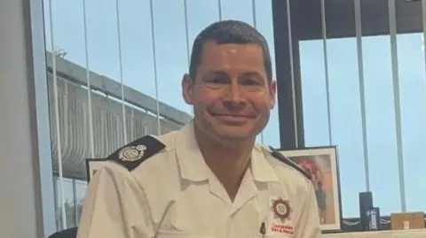 Joe Haustead smiling at the camera. He is wearing uniform. Behind him is a window with blinds and a view of blue skies.