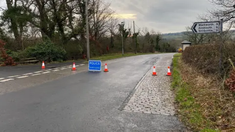 Jon Douglas A rural road with cobbles on on side and a large blue sign from the police saying road closed 