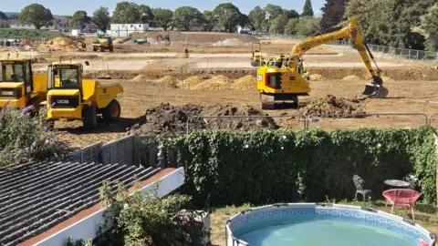 Kayleigh Paterson A garden with a paddling pool that backs onto a construction site. There are yellow diggers on the site. Trees are surrounding the area and there are some houses in the distance.