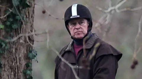 Andrew in a horse riding hat that is black with a white stripe and wearing a brown jacket.
