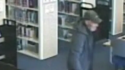 A blurred CCTV image of a man who is walking through the library. He is wearing a black hooded coat and a dark grey or green cap. There are a number of library shelves behind him, as well as a library table. 