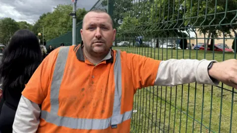 A man in an orange work polo with grey stripes stands next to a wire fence.