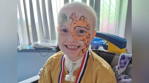 An eight-year-old girl wearing a yellow top is sitting in a hospital room with a small plastic tube and device attached to the front of her neck. She is smiling, has no hair, and there is an orange giraffe image painted on the right side of her face.