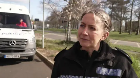 Liz is stood outside on an open pathway in her uniform being interviewed. A van is parked close by with a man inside