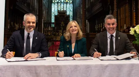 West Yorkshire Combined Authority Image shows three people signing an agreement at table with a white tablecloth on it. From left to right is Oliver Coppard Mayor of South Yorkshire in a blue suit and blue tie with white shirt; Tracy Brabin is in centre shot with bobbed blonde hair and a green jacket; David Skaith is to the right of shot in a grey suit with black tie. There is a vase of white roses on the table. 