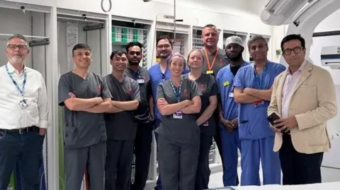 East Kent Hospitals NHS Trust The team at the Kent and Canterbury Hospital in Canterbury standing in the dedicated stroke thrombectomy suite. Eleven people, nine of whom are in scrubs, are in the picture. At either end are men in formal clothes.