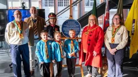 Claire Louise Photography Lord-Lieutenant of Tyne & Wear Lucy Winskell OBE, in a uniform is standing next to the Blue Plaque with Ben Hodgkiss, UK Contingent Leader for the Scouts and Sunderland Mayor Allison Chisnall. Three young scout memebrs in blue shirts and yellow and red scarves are smiling in front of the plaque. 